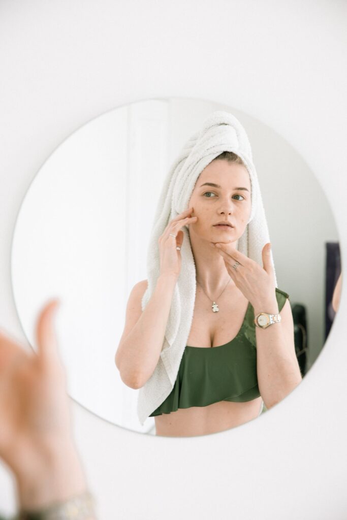 Young woman examining her facial skin in the mirror - acne scars and uneven skin texture